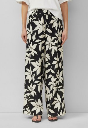 Czarne spodnie o szerokich nogawkach z dużym białym kwiatowym wzorem, noszone z białym topem i czarnymi sandałami z odkrytymi palcami, stojące na jednolitym tle.