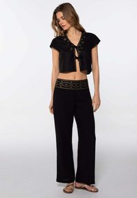 Top corto negro con tachuelas decorativas y un lazo en la parte delantera, combinado con pantalones negros de pierna ancha que presentan una cinturilla adornada. Textura suave.