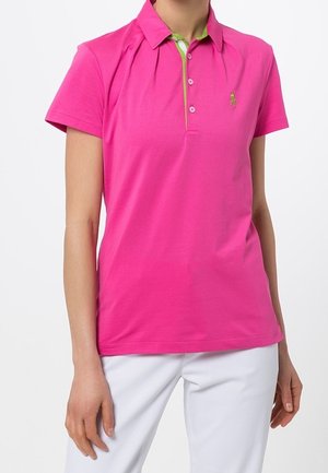 Vrouw draagt een felroze polo met korte mouwen en witte knopen en een witte broek tegen een effen achtergrond.