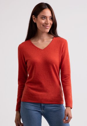 Roter Pullover mit langen Ärmeln und V-Ausschnitt. Weiche Textur, taillierte Form und subtiler Melangeeffekt. Kombiniert mit blauen Jeans.