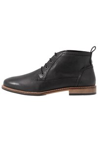 Botín negro de cuero hasta el tobillo con punta redonda, diseño de cordones, suela marrón en contraste y detalles de costura en el lateral. Cuenta con una lengüeta.
