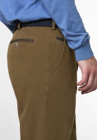 Close-up of a person wearing brown pants with back pockets and a black woven belt, dressed in a blue long-sleeve shirt.