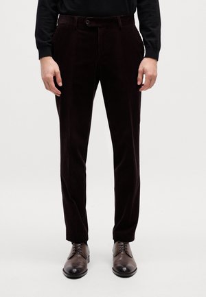 Homme portant un pantalon en velours côtelé marron foncé avec bouton et fermeture éclair, accompagné de chaussures habillées marron foncé à lacets, debout sur un fond clair.