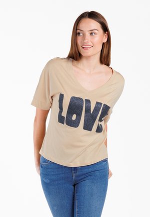 Camiseta de cuello en V beige con mangas cortas que presenta un gran gráfico negro de "LOVE". Tejido suave con un ajuste relajado, combinada con jeans azules.