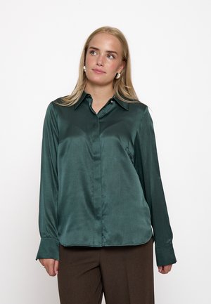 Femme aux longs cheveux blonds portant un chemisier en satin vert foncé et un pantalon marron, regardant légèrement sur le côté sur un fond blanc.