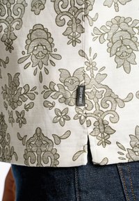 Tela beige con intrincados patrones florales verdes, que presenta una etiqueta en la costura lateral y un fondo de denim oscuro en contraste.