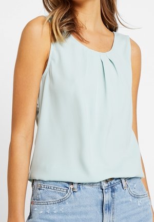 Bezrękawna jasnoniebieska bluzka z okrągłym dekoltem i zakładką z przodu, noszona wpuszczona w wyblakłe niebieskie dżinsy.