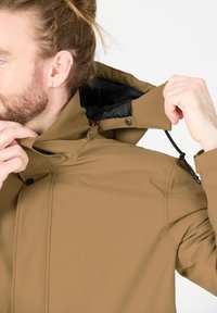 Bruine waterdichte jas met een hoge kraag, interne zwarte voering, drukknoopsluitingen en een verstelbaar trekkoord bij de hals.