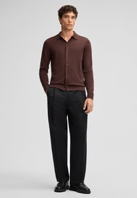 Camicia marrone a maniche lunghe con colletto, texture liscia e lucentezza sottile, abbinata a pantaloni neri plissettati e scarpe nere.