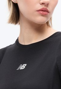 Γυναίκα που φοράει μαύρο μπλουζάκι New Balance, εμφανίζεται το κάτω μέρος του προσώπου της, ο λαιμός και μέρος κοντών ξανθών μαλλιών με ένα μικρό στρογγυλό σκουλαρίκι.