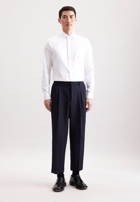 Camicia bianca con colletto alto, abbinata a pantaloni tailleur blu scuro. Il modello indossa scarpe stringate nere, in piedi su uno sfondo chiaro.