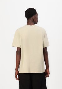 T-shirt beige à manches courtes en coton, avec un col rond, une coupe décontractée et une texture lisse. La vue arrière montre des détails minimaux.