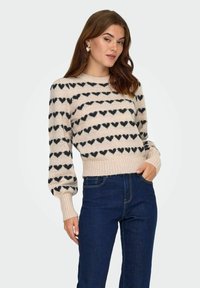 Suéter de punto beige con un patrón de corazones negros, que cuenta con un escote redondo, mangas abullonadas y dobladillo acanalado. Combinado con vaqueros oscuros de mezclilla.