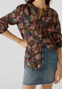 Femme portant un chemisier long à manches coloré avec des boutons noirs et une jupe en denim bleu avec des poches avant et un ourlet effiloché.