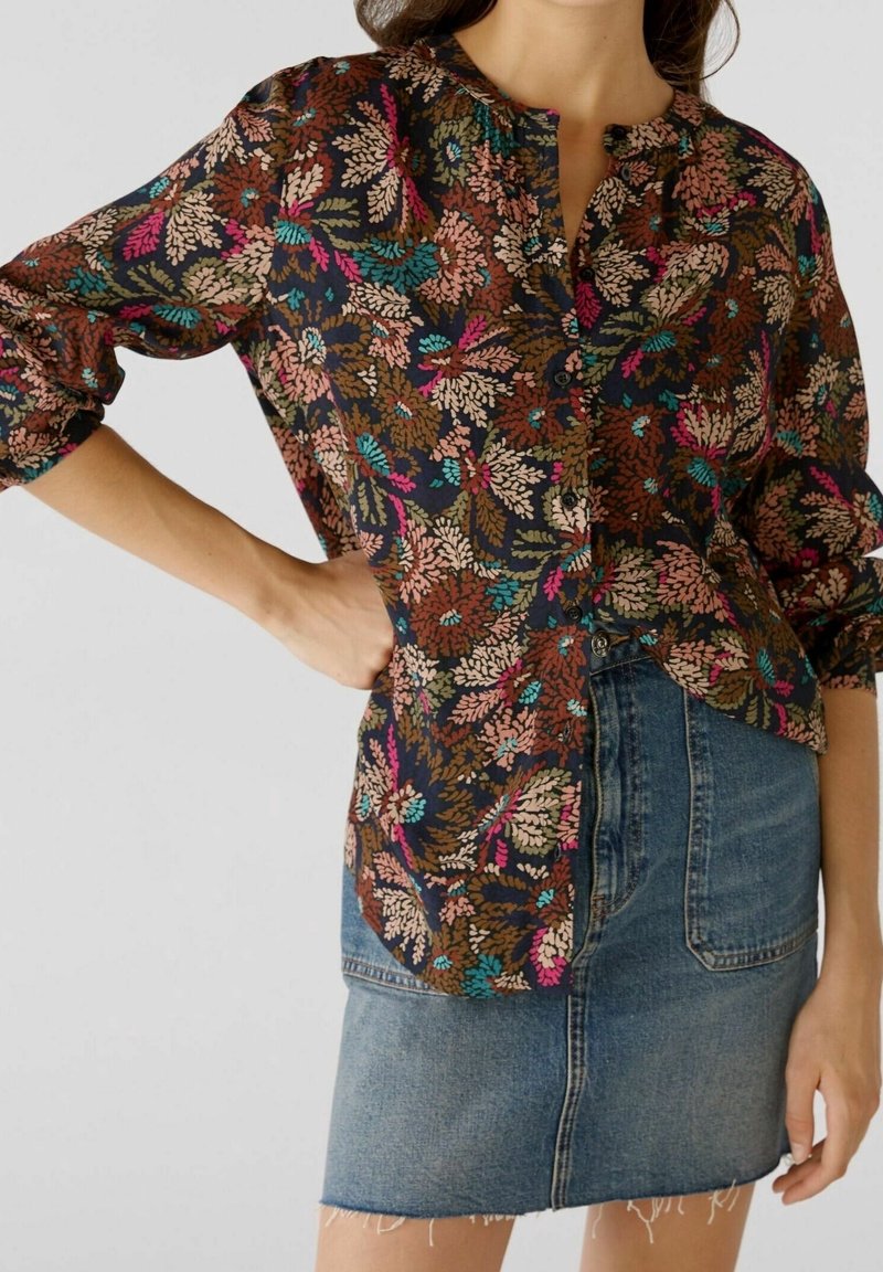 Femme portant un chemisier long à manches coloré avec des boutons noirs et une jupe en denim bleu avec des poches avant et un ourlet effiloché.