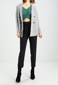 Donna che indossa un crop top verde a balze, un blazer doppiopetto grigio a scacchi, pantaloni neri a vita alta con fiocco e stivaletti neri.