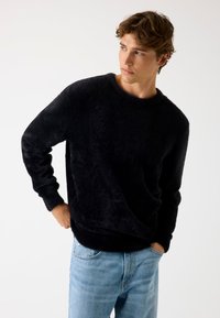 Zwarte, fuzzy trui met een ronde hals en lange mouwen, gecombineerd met lichtblauwe jeans. De stof heeft een zachte, gestructureerde uitstraling.