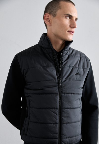 Black puffer vest with a high collar and zip front, featuring horizontal quilting and side pockets. Worn over a black long-sleeve shirt.