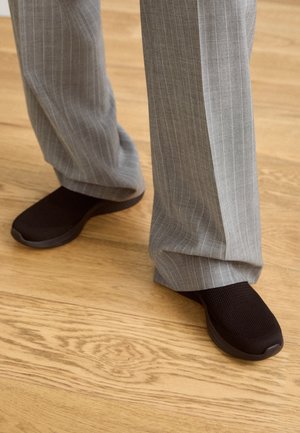 Des chaussures noire à enfiler avec un dessus texturé, associées à un pantalon à rayures fines gris clair, reposant sur un sol en bois.