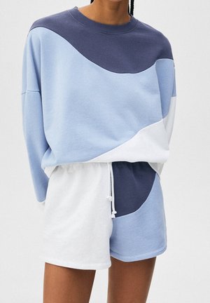 Person iført sweatshirt og matchende shorts i lyseblå, marineblå og hvid i blokfarver med snøre i taljen mod en ensfarvet baggrund.