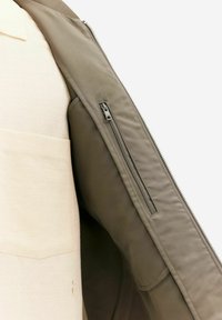Olive green jacket with a smooth texture, featuring a zippered inner pocket on the left side and a beige, textured inner shirt.