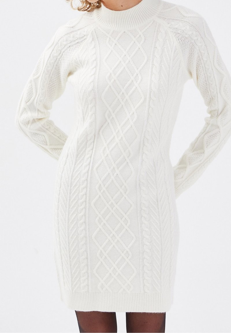 Femme portant une robe pull en maille blanche avec un col montant et des manches longues, se tenant les mains sur les hanches contre un fond blanc.