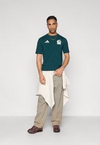 Zöld rövid ujjú ing logóval és fehér csíkokkal, bézs nadrág és barna sportcipő. Deréknál megkötött réteges világos ing.