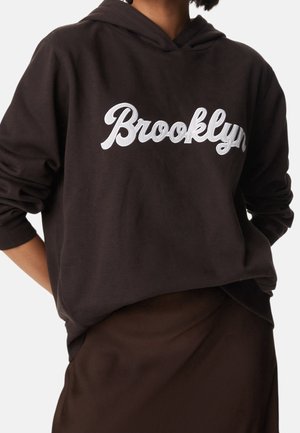 Osoba ubrana w ciemnobrązową bluzę z kapturem z dużym białym napisem "Brooklyn" na piersi oraz dopasowaną ciemnobrązową spódnicę lub spodnie.