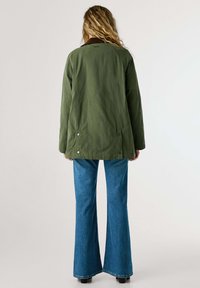 Giacca verde oliva con colletto marrone, caratterizzata da una vestibilità rilassata e chiusure a scatto sui lati, abbinata a jeans a zampa d'elefante blu.