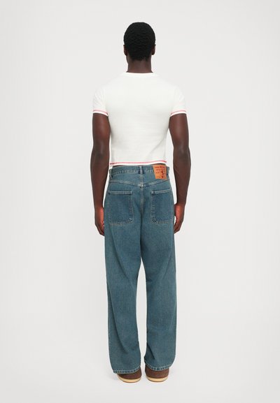 Man in een wit, aansluitend T-shirt met korte mouwen en ruime blauwe jeans, staand voor een vlakke witte achtergrond met de handen ontspannen langs de zijkanten.