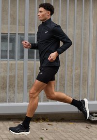 Black running jacket with a high collar, paired with black shorts. The runner wears black socks and shoes with white soles, in motion on a walkway.