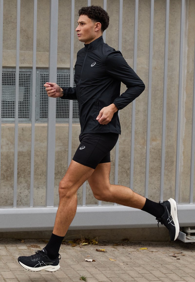 Chaqueta de running negra con cuello alto, combinada con pantalones cortos negros. El corredor lleva calcetines negros y zapatillas con suelas blancas, en movimiento sobre un sendero.