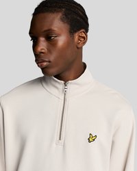 Jeune homme avec des cheveux tressés portant un sweat-shirt beige à fermeture éclair quart avec un petit logo d'oiseau jaune sur la poitrine.