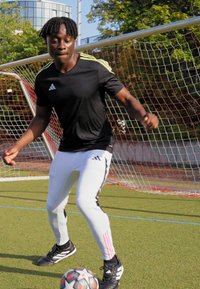 Schwarzes Sportoberteil mit grünen Akzenten, weiße enganliegende Hose mit schwarzen und pinken Streifen sowie schwarze Fußballschuhe auf einem Rasenplatz.