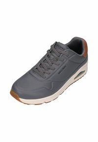 Zapatilla sintética gris con detalles perforados, cordones planos y acentos marrones en el talón. Mediasuela blanca y suela de goma texturizada.
