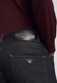 Person som bär mörka denimjeans med orange sömmar och Emporio Armani-logotyp för patch och metall-emblem på bakfickan, matchat med en vinröd tröja.