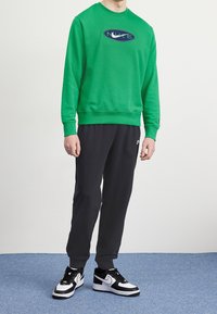 Grön sweatshirt med blå logga, kombinerad med svarta sweatpants och svartvita sneakers, visad mot en neutral bakgrund.