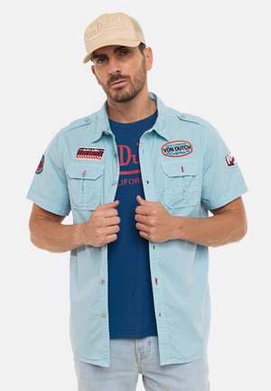 Homme portant une casquette beige Von Dutch, une chemise à manches courtes bleu clair avec des patchs, et un T-shirt bleu marine Von Dutch, posant avec les mains sur la chemise.