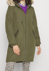 Parka verde oliva con capucha de piel sintética, cremallera frontal, cintura ajustable con cordón y dos bolsillos con cremallera, combinada con leggings estampados.