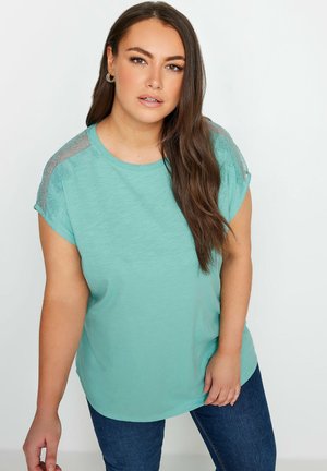 Lys teal kortærmet bluse med rund halsudskæring og gennemsigtige blonde detaljer på skuldrene, parret med mørkeblå jeans. Glat tekstur.