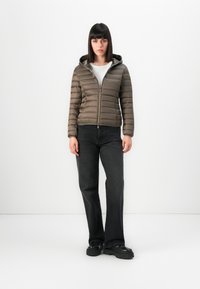 Vrouw in een bruine gewatteerde jas met capuchon, witte top, zwarte jeans en zwarte chunky schoenen, staand tegen een effen witte achtergrond.
