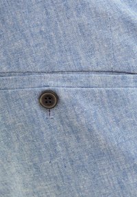 Hellblauer Stoff mit strukturierter Oberfläche und subtilen Variationen. Verfügt über einen schwarzen Vierlochknopf und eine gestickte Taschenapplikation.