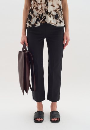 Pantaloni neri su misura con una vestibilità slim, abbinati a una blusa a fantasia nei toni beige e marroni e sandali neri scivolati. Si tiene una borsa a tracolla in pelle marrone.