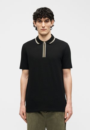 Νεαρός με σκούρα, σγουρά μαλλιά φοράει μαύρο πόλο μπλουζάκι με κοντά μανίκια και ριγέ γιακά με φερμουάρ, στέκεται μπροστά σε απλό φόντο.