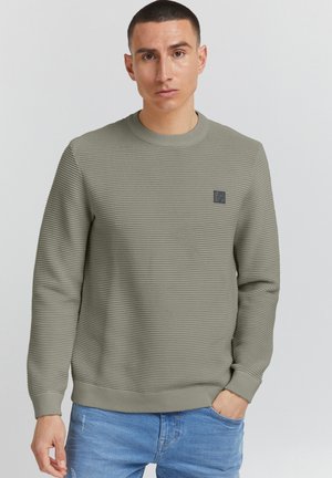Olive green ribbed sweater with a crew neck, long sleeves, and a small textured logo patch on the chest. Material appears soft and textured.
