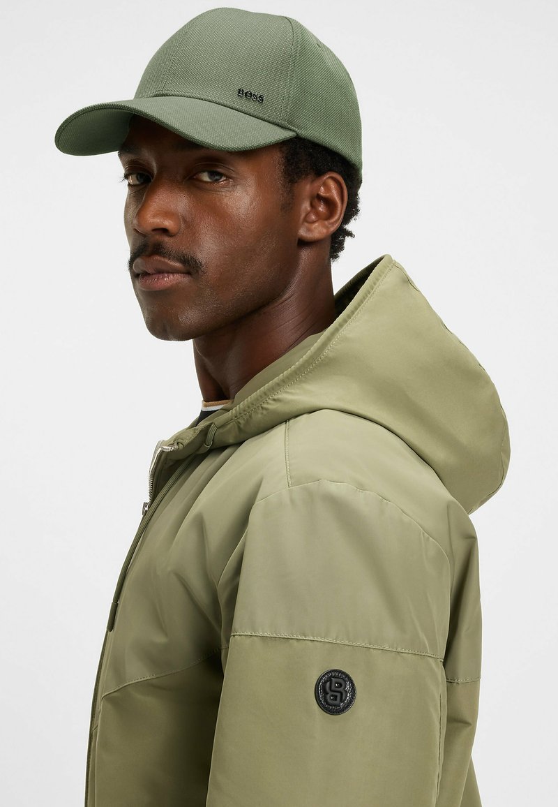 Green baseball cap with textured fabric and embroidered logo, paired with a matching olive green hooded jacket featuring a logo patch.