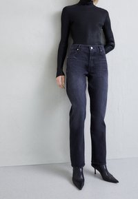 Musta ribattu korkea kaulus yhdistettynä tummanpesuisiin suoriin farkkuihin. Farkuissa on tavalliset taskut ja helma, jossa on hieman rispaantumista. Mustat kärjelliset korkokengät.