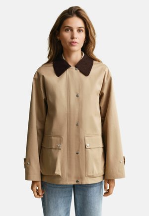 Frau trägt eine beige Jacke mit einem dunkelbraunen Kragen, vorderen Druckknöpfen, zwei vorderen Klappentaschen und blauen Jeans, steht vor neutralem Hintergrund.