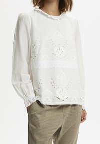 Vit broderad blus med intrikata snitt, en scalloped halsringning och elastiska ärmslut. Matchad med rutiga byxor.