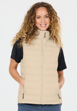 Lächelnde Frau mit lockigem Haar, die eine beige, gesteppte, ärmellose Weste über einem schwarzen Kurzarmshirt trägt, die Hände in den Westentaschen.